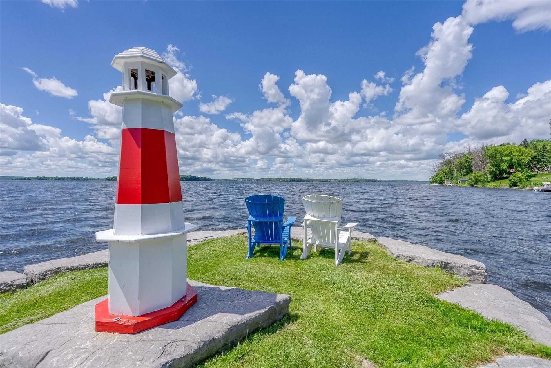 Red-and-white lighthouse by a lakeshore with two blue chairs under a cloudy sky.