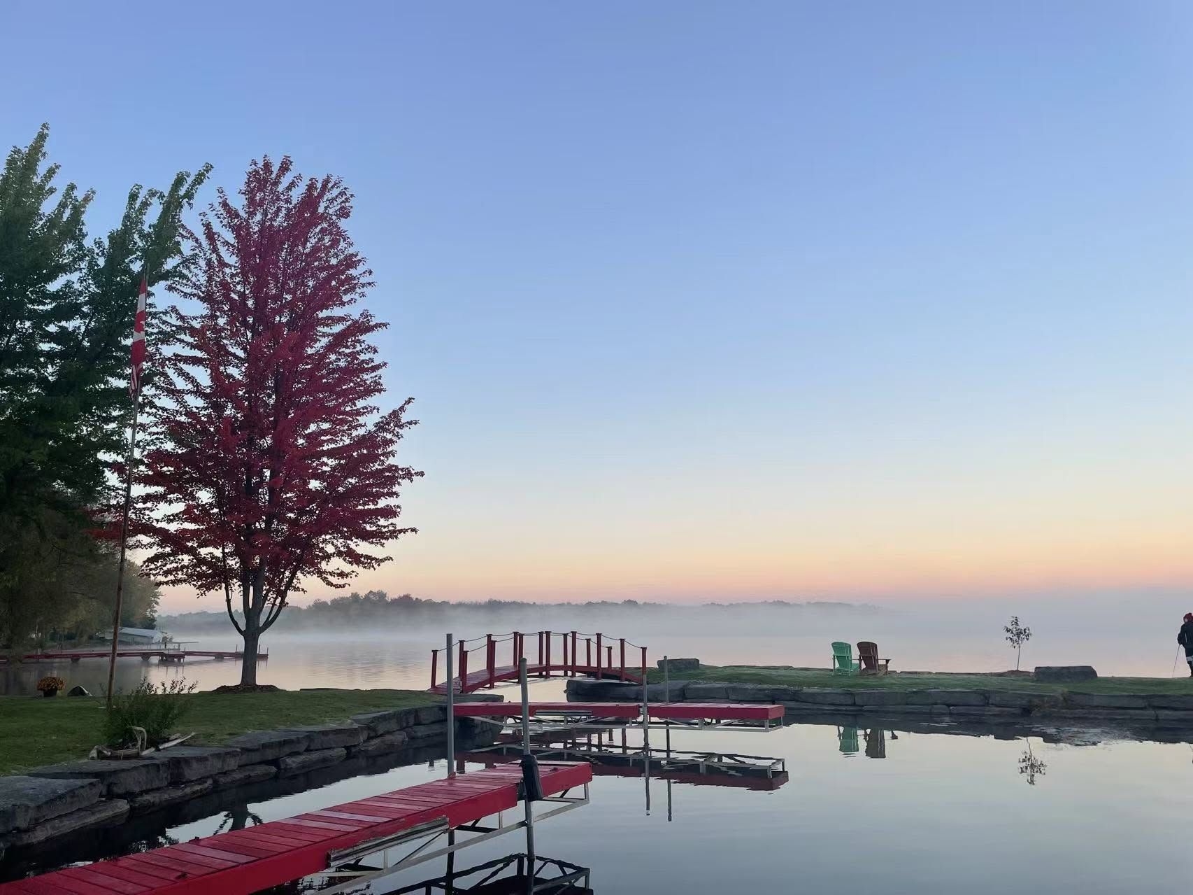 Calm lakeside dock at sunrise with red kayaks, a tree, and mist over still water