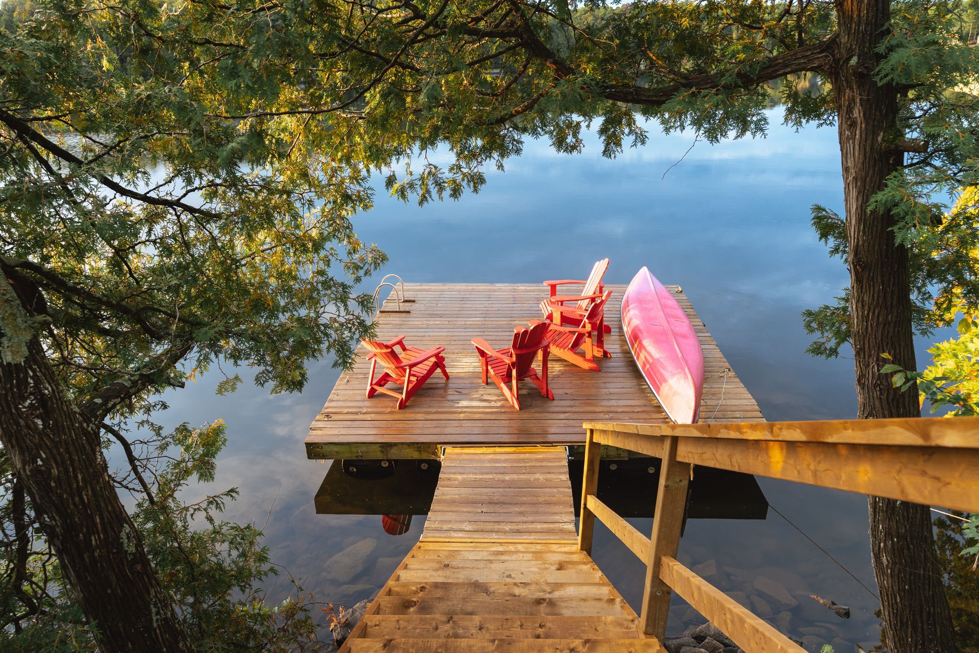 Wooden dock with red chairs and pink kayak beside a calm lake, framed by trees