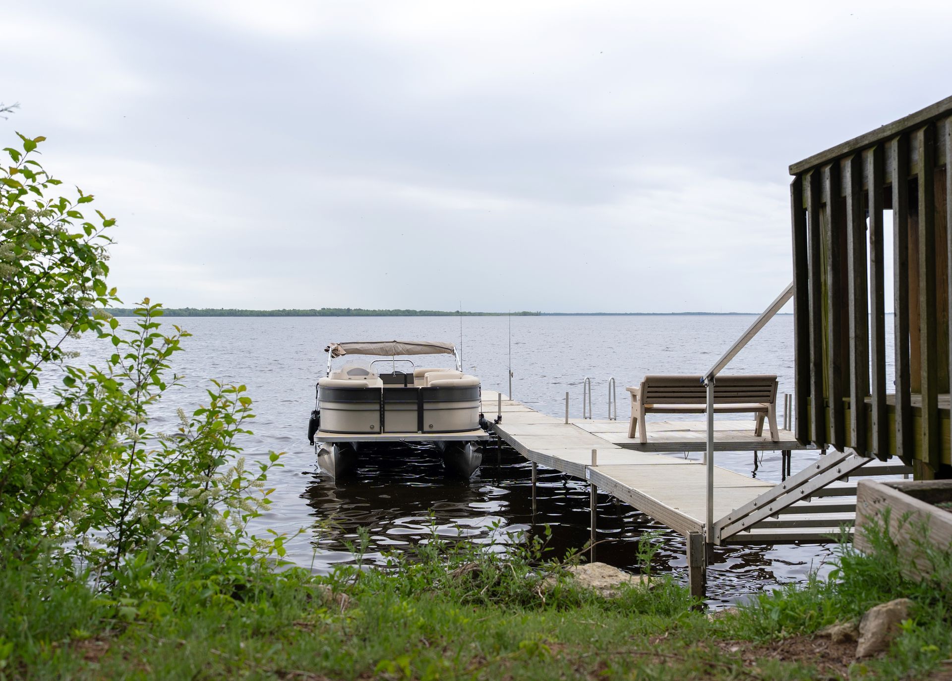 Wooden dock with chairs and a boat on a calm lake, framed by greenery and a cloudy sky