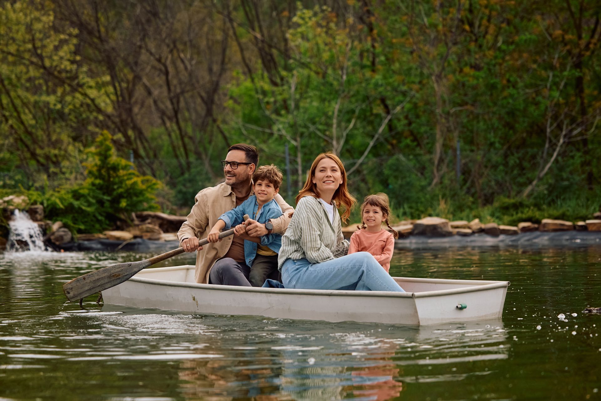 Family in a small rowboat on a calm river, with trees and a waterfall in the background.
