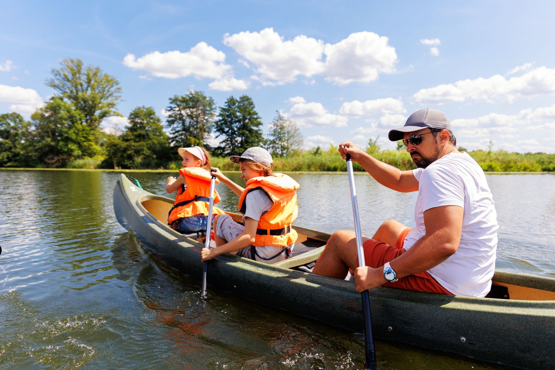 Three people canoeing on a calm lake on a sunny day, with trees and clouds in the background.