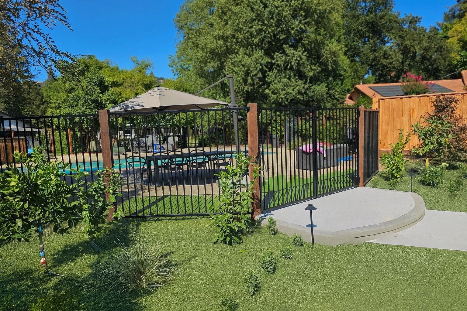 Black metal fence surrounds a backyard pool. A wooden gate leads to a patio with an umbrella and table.