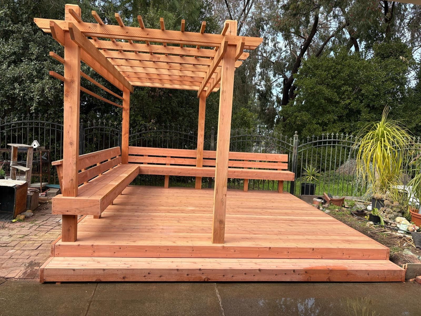 Wooden pergola with built-in benches on a deck in a backyard, surrounded by trees.