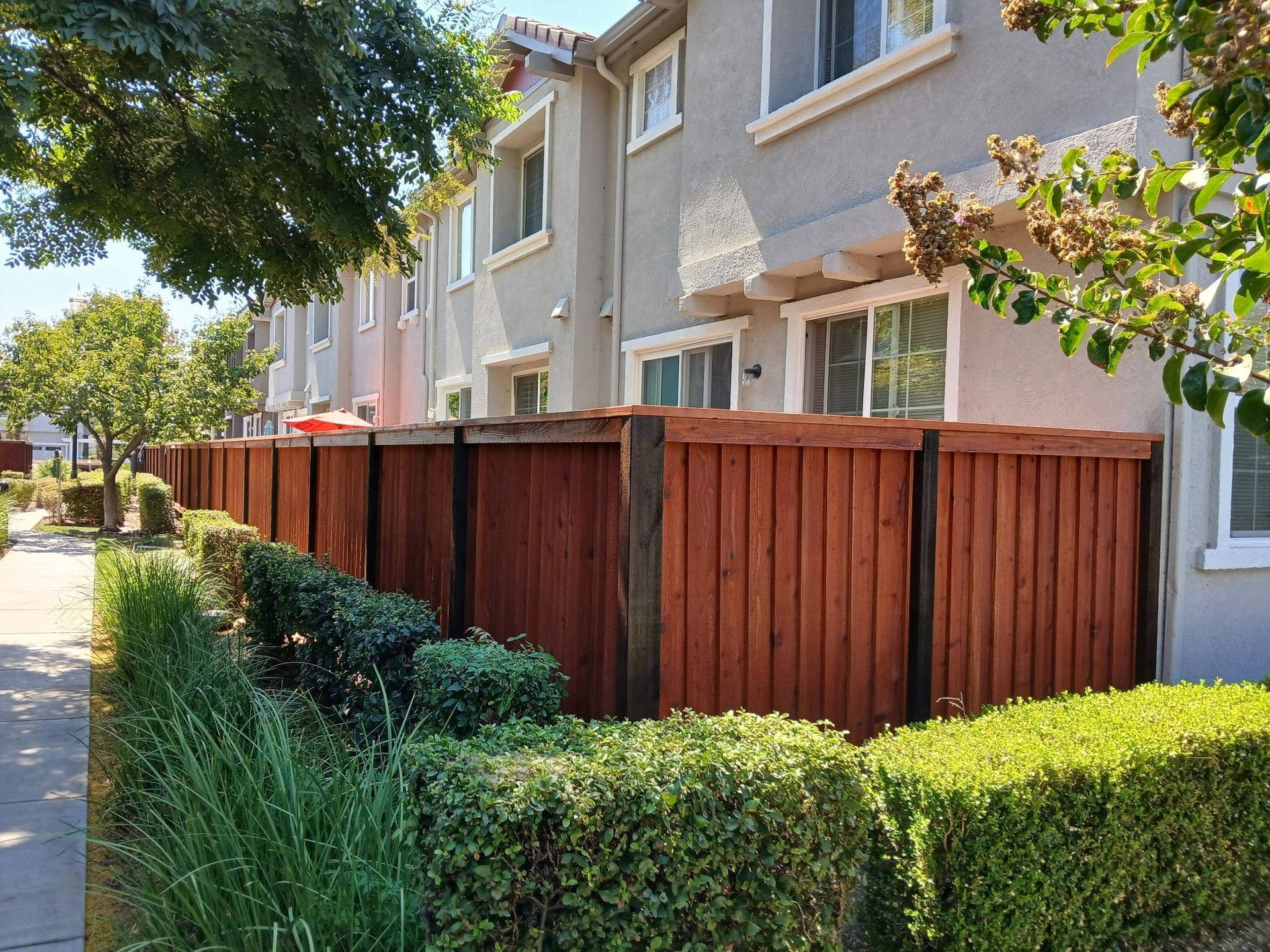 Row of townhouses with a stained wooden fence and green shrubbery along a sidewalk.