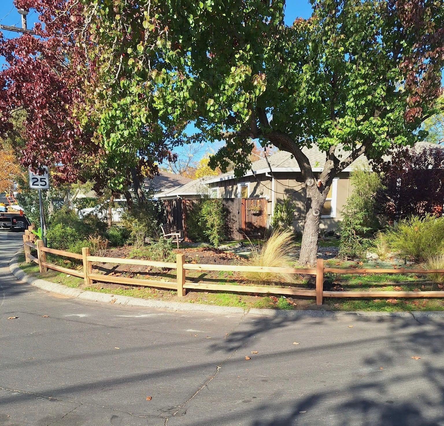 A split-rail fence lines a curved street, surrounding a house with fall trees and a speed limit sign.