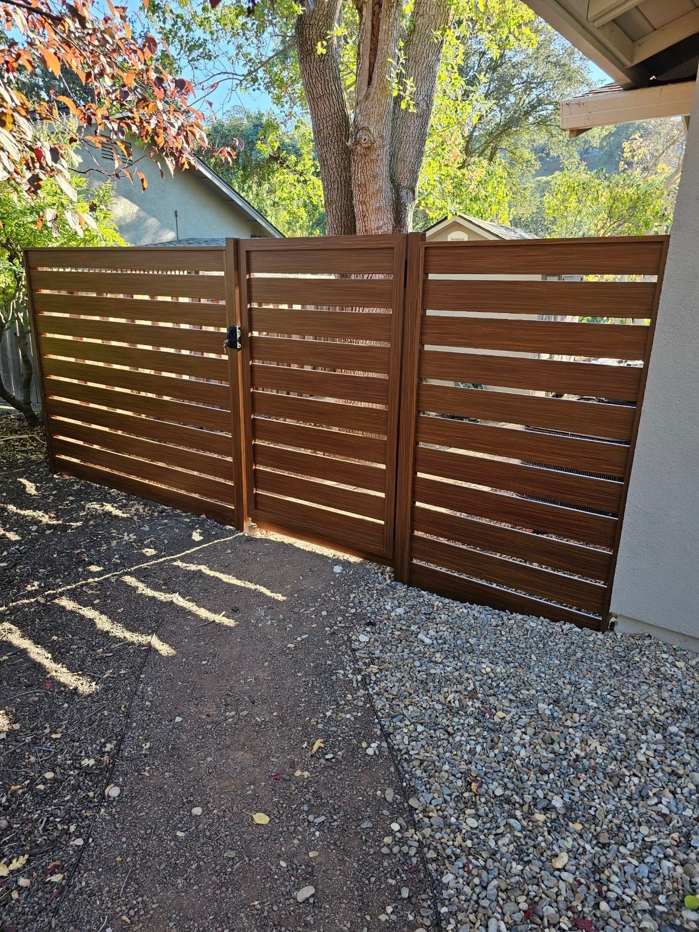 Brown horizontal slat fence with gate, gravel ground, tree in background.