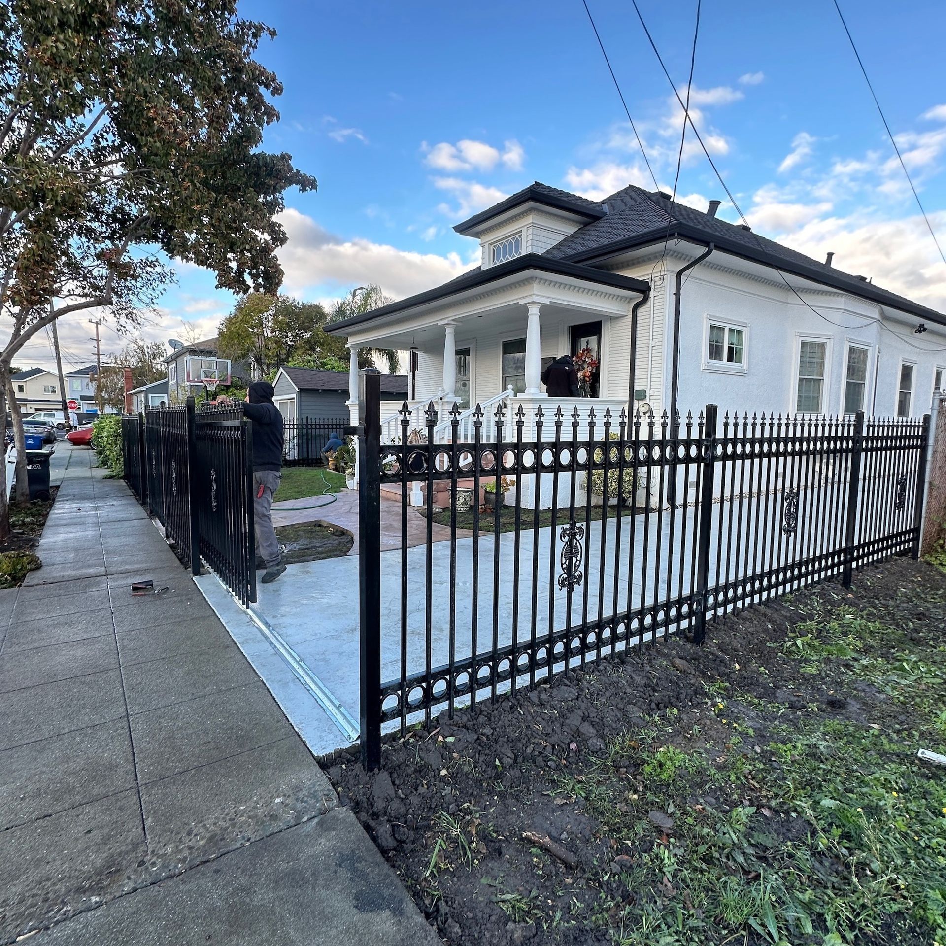 White house with black wrought iron fence alongside a sidewalk under a partly cloudy sky.