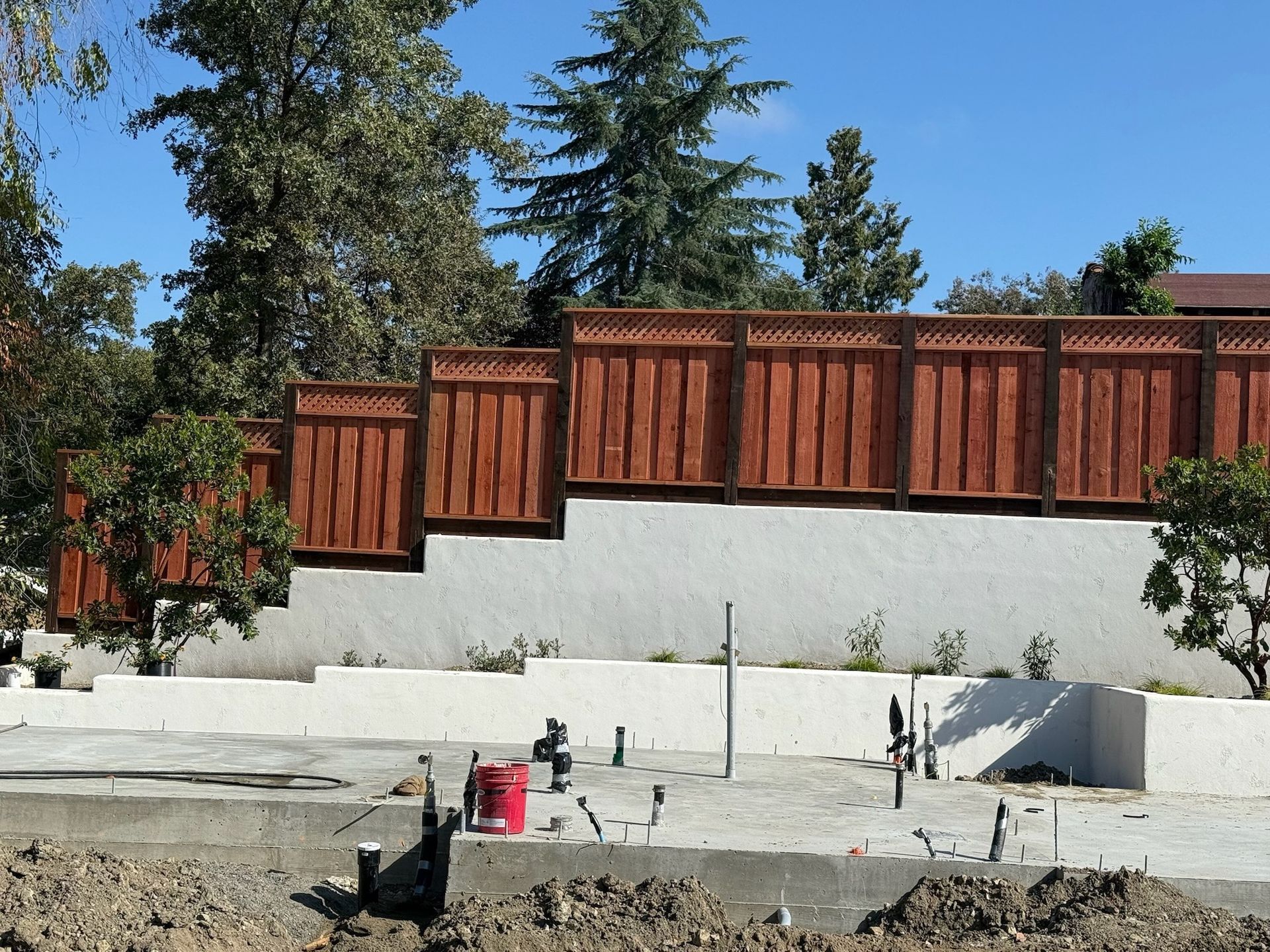 Wooden fence atop a stucco wall, with construction in foreground and trees/sky in background.