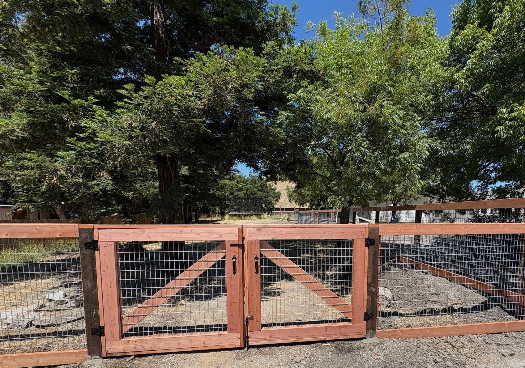 Wooden gate in a fence, with trees and a house in the background on a sunny day.