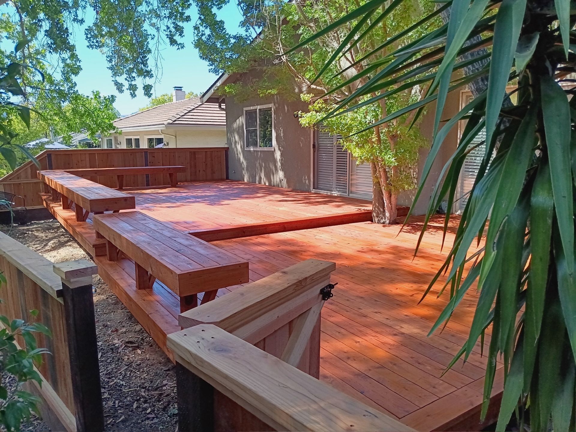 Wooden deck with built-in benches, stained reddish-brown, next to a small building and greenery.