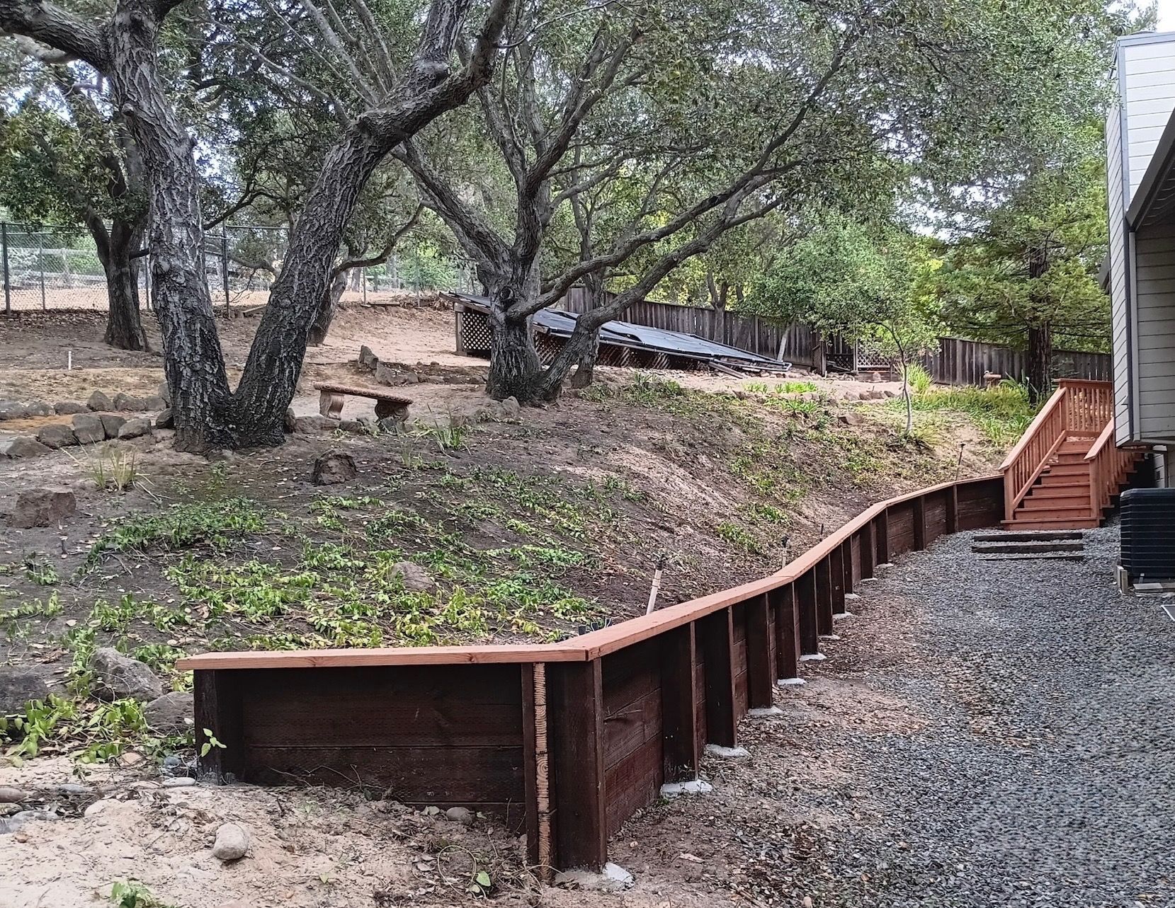 Wooden retaining wall and stairs in a backyard with trees and a house.