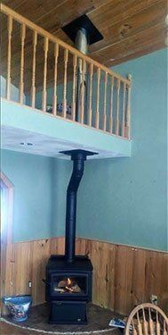 Wood-burning stove with black pipe extending upwards. Balcony with wooden railing above, teal wall and wooden paneling.