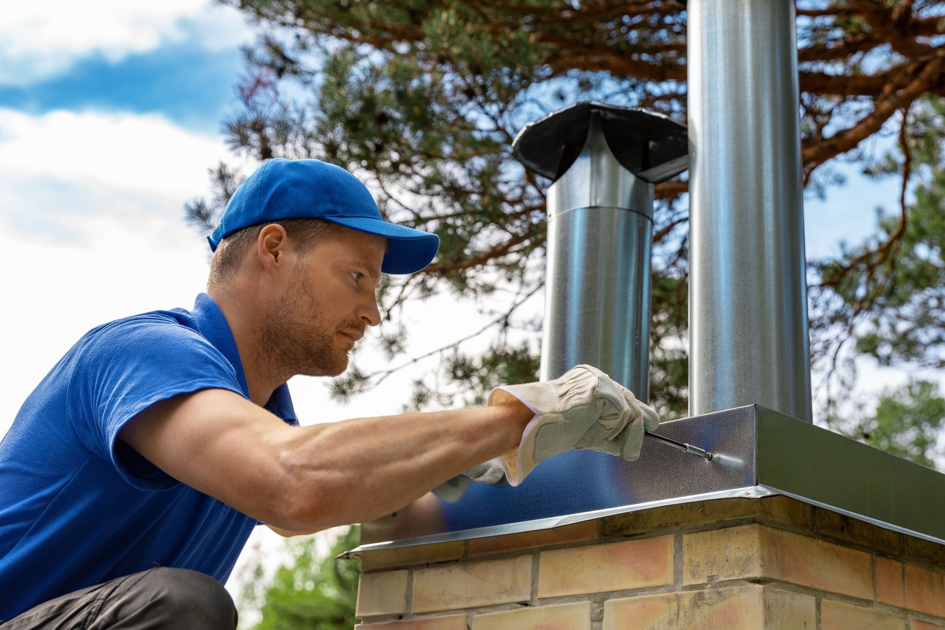 Man in blue cap and shirt working on a chimney top, wearing gloves.