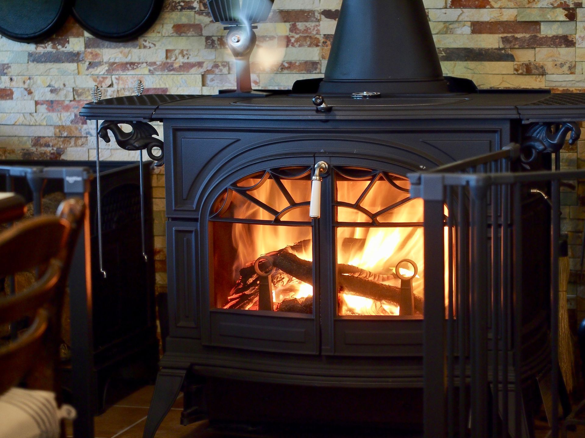 A wood-burning stove with flames inside. It's black, and the brick wall behind it has various pots hanging.