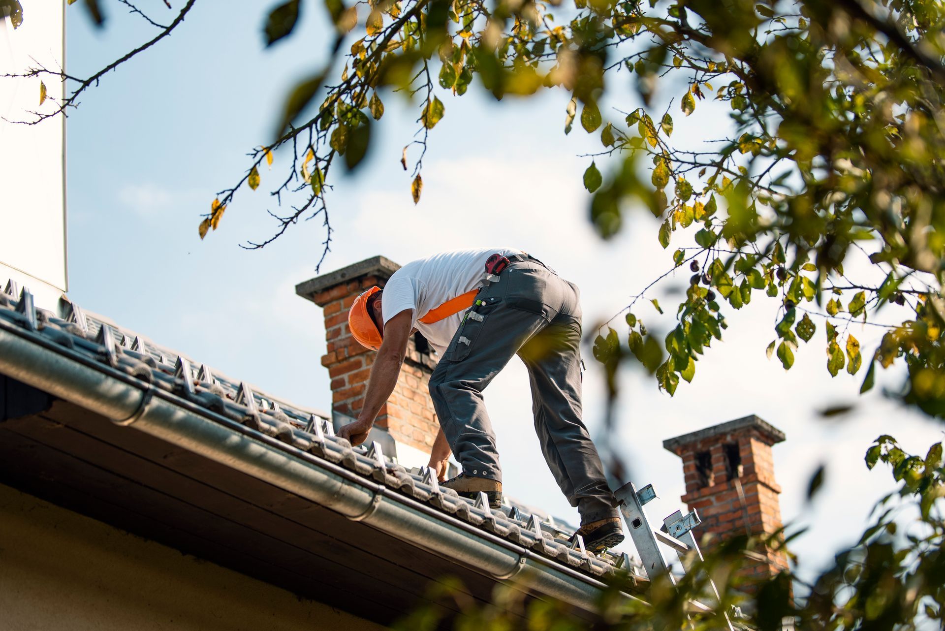 Roofer on ladder near chimney, fixing roof. He wears a hard hat and orange shirt. Sunny day.