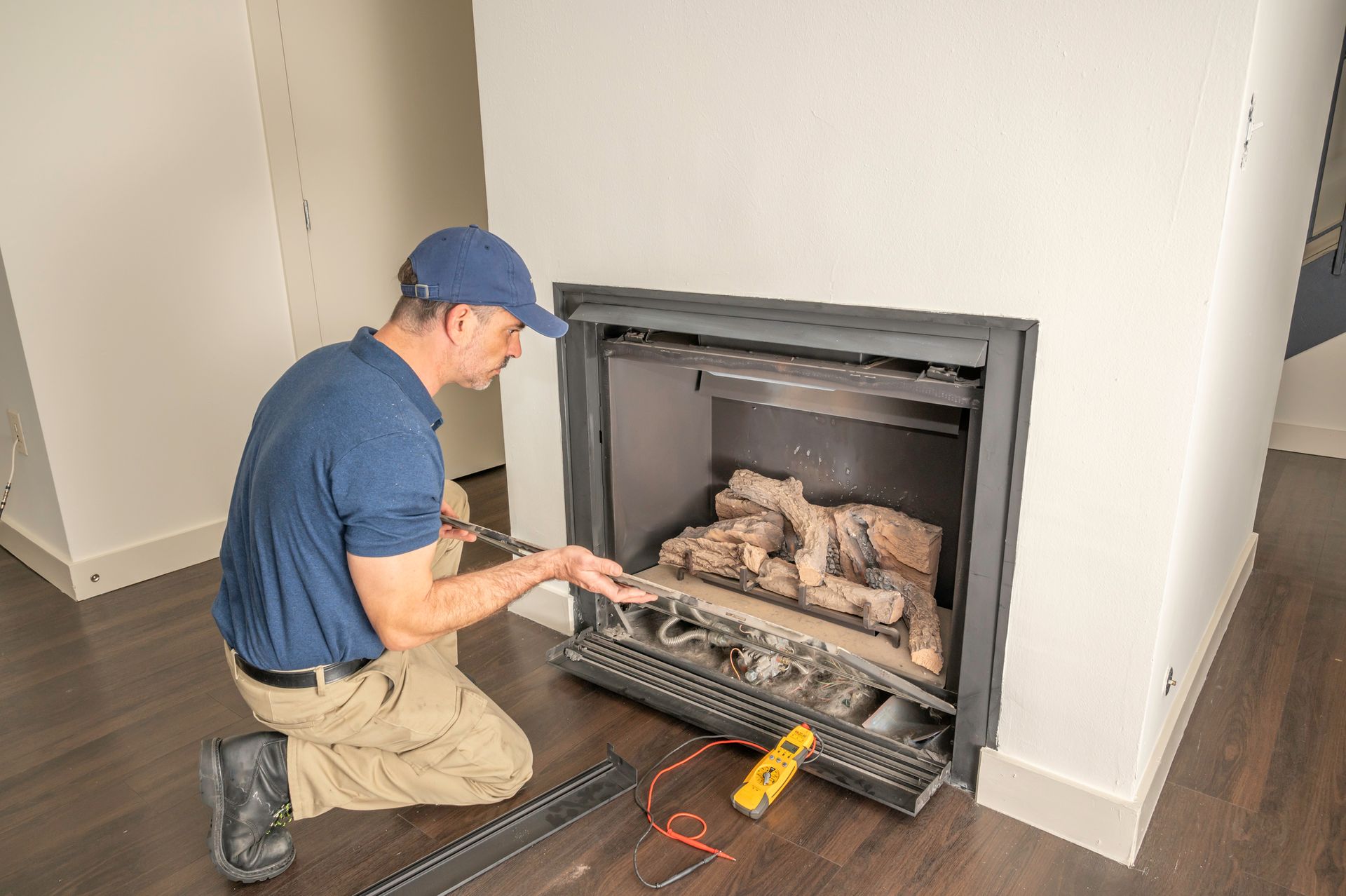 Man in blue shirt kneels to inspect a fireplace, with tools on floor. Interior setting, neutral colors.