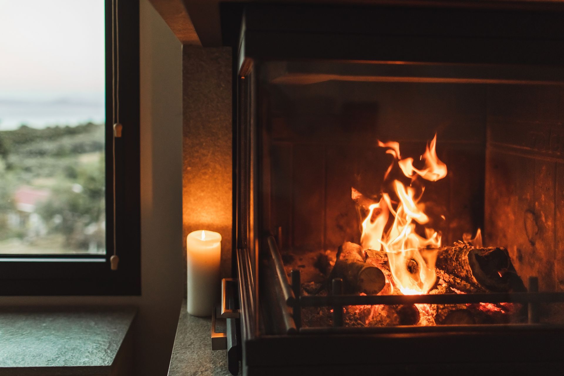 Fireplace with burning logs, illuminated by flames. A candle glows beside the window with a scenic view.