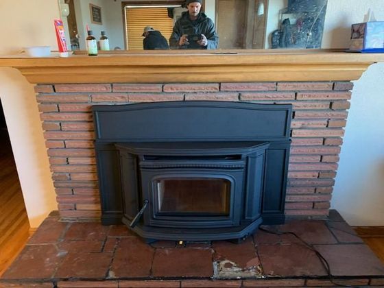 A black pellet stove installed in a red brick fireplace. A person is reflected in the mirror above.