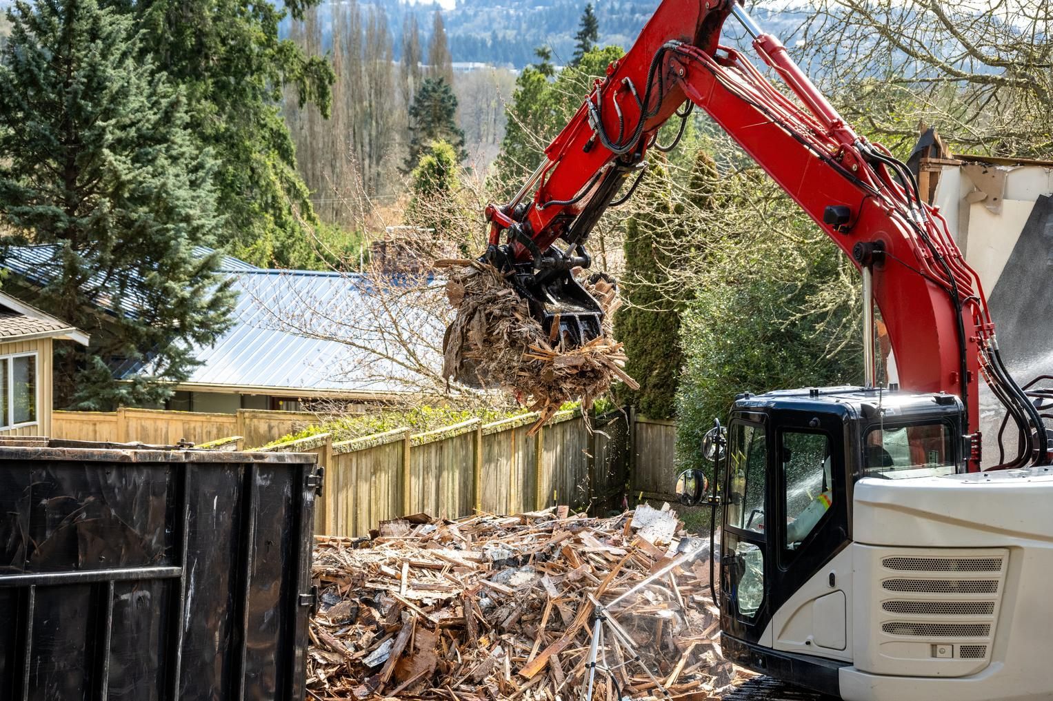 A red excavator is loading a pile of wood into a dumpster.