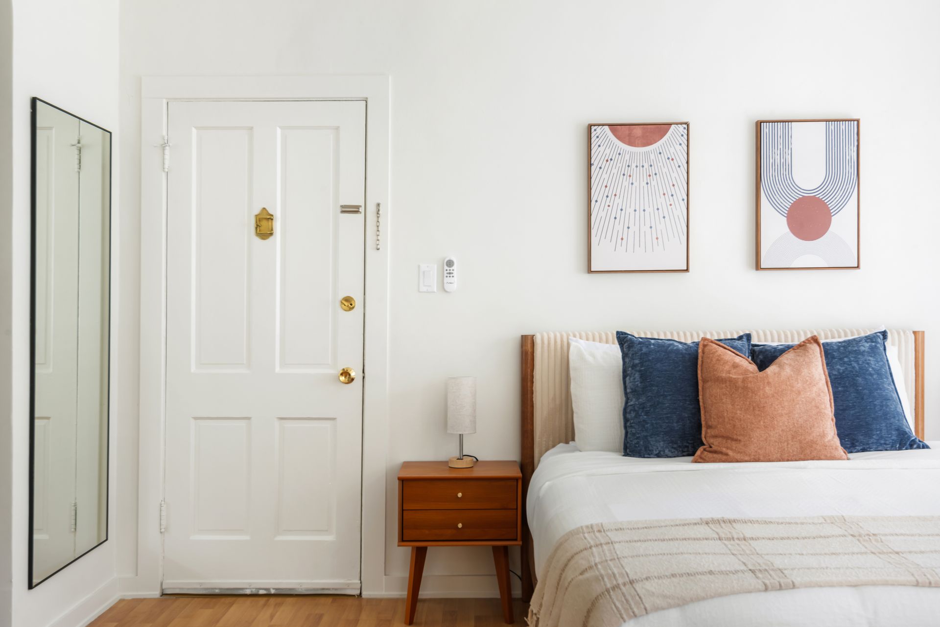 Bedroom with a white door, framed mirror, wooden nightstand, bed with pillows, and artwork.