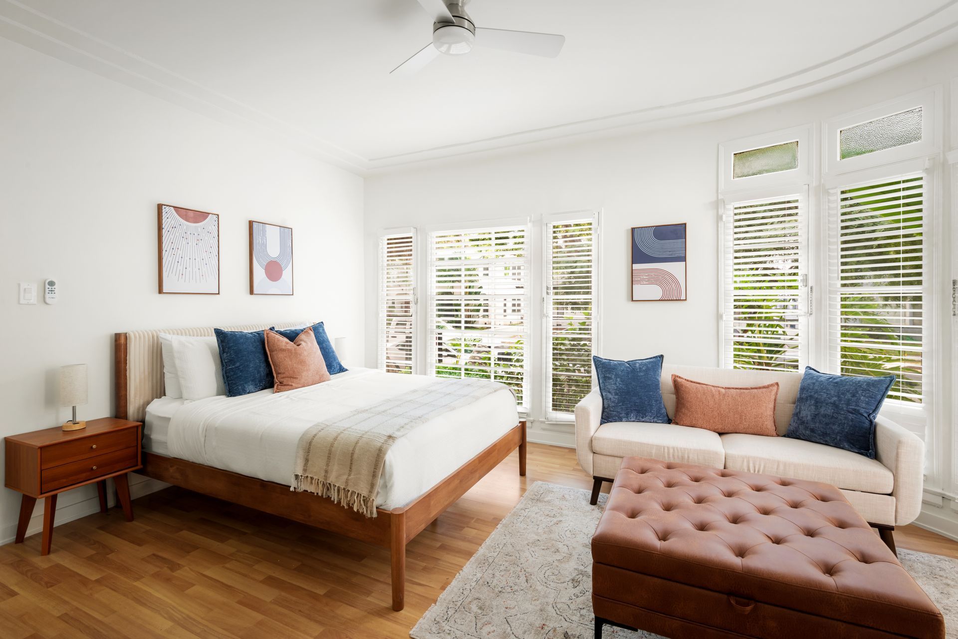 Bedroom with a bed, sofa, and ottoman; white walls, wooden floor, large windows, and art.
