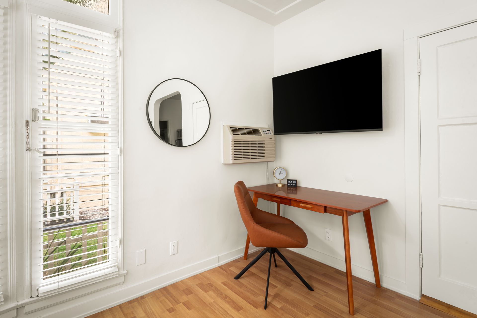 Small home office with desk, chair, window, TV, mirror, and white walls.