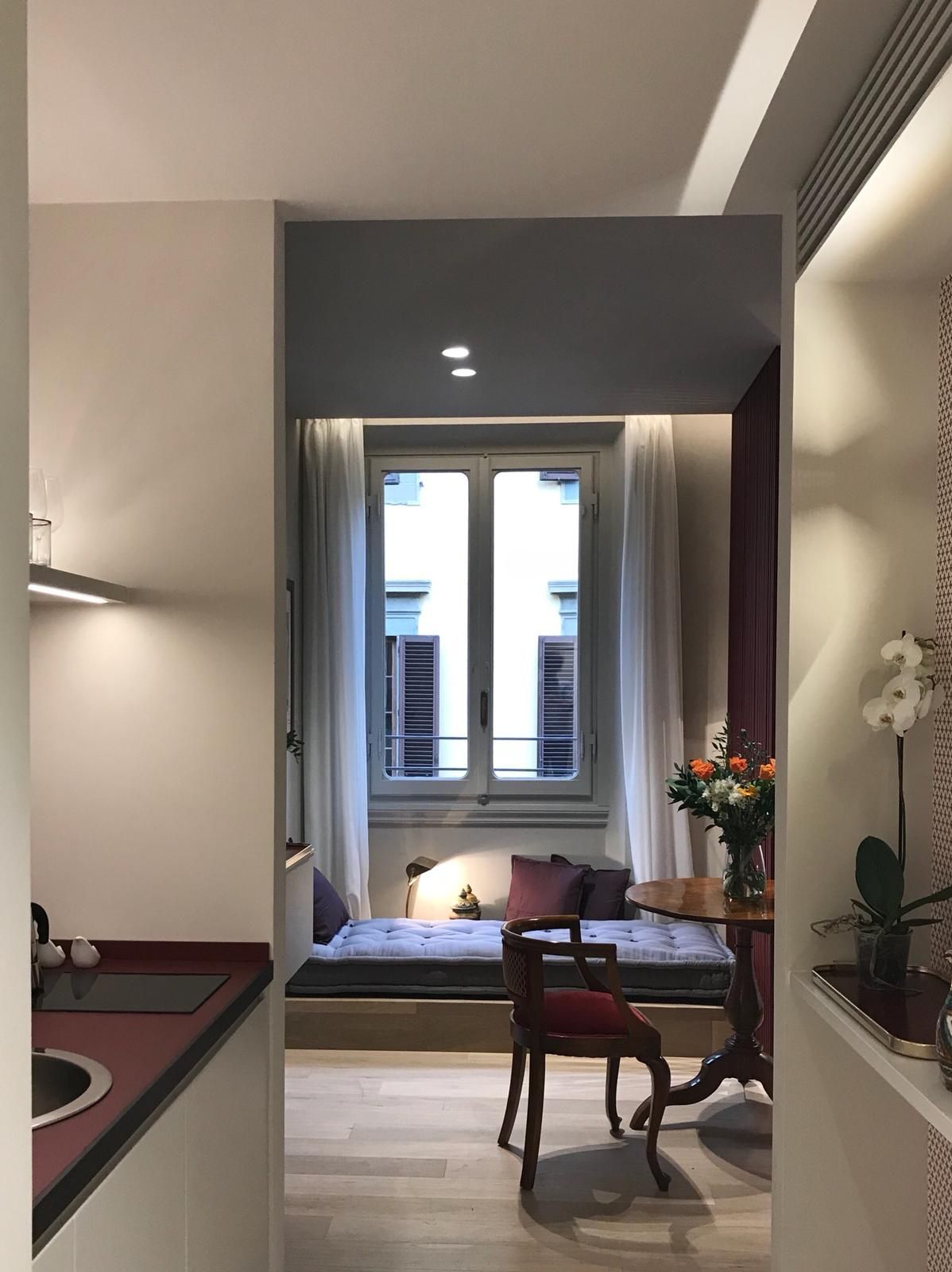 Accogliente angolo con panca vicino alla finestra, tavolo e sedia. Interni rossi e bianchi con fiori, tende e illuminazione.