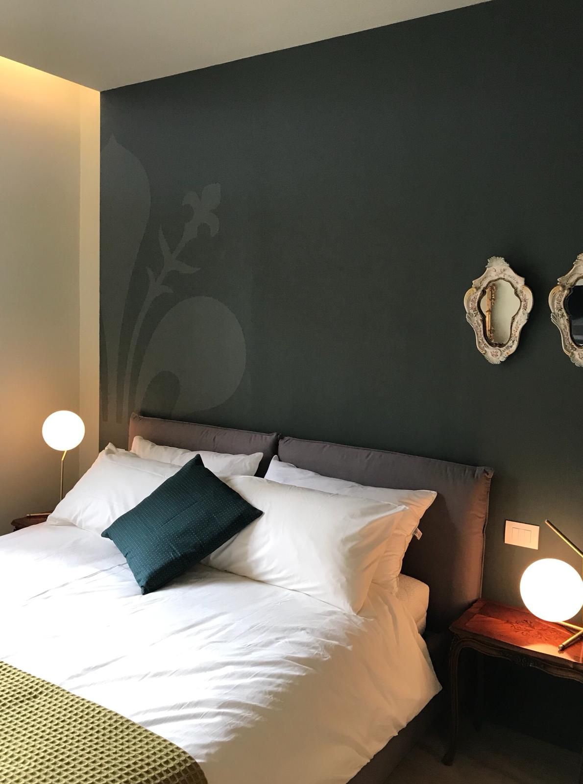 Camera da letto con letto, parete decorativa verde scuro, piccoli tavolini laterali e specchi decorativi.