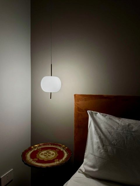 Una lampada a sospensione illumina il comodino e la testiera. Sono visibili un cuscino bianco e un vassoio decorato.