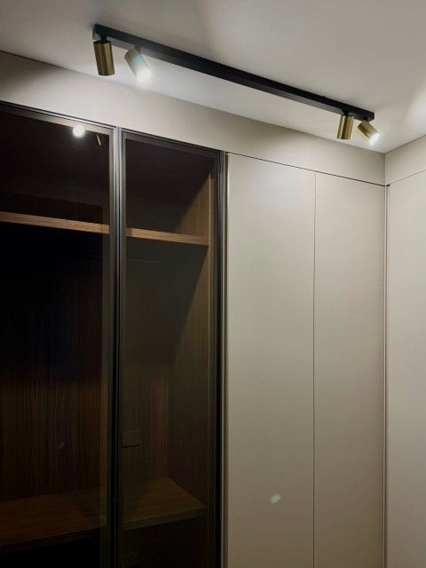 Un binario di illuminazione nero illumina un armadio a muro con ripiani in vetro e legno e ante beige chiaro.