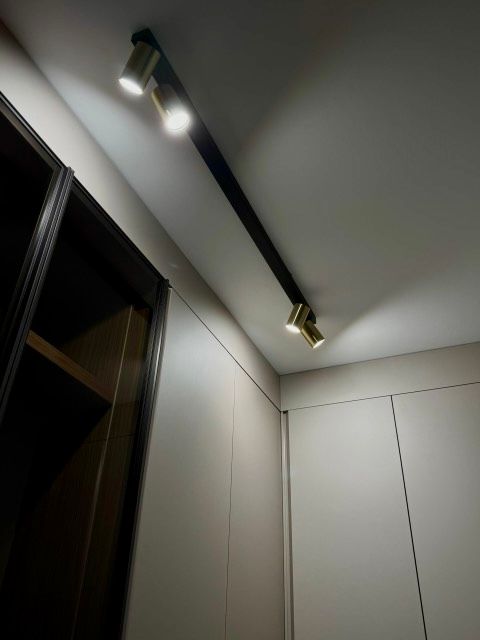 Un binario di illuminazione nero con tre faretti dorati illumina un soffitto bianco e le pareti vicino a un mobile scuro.