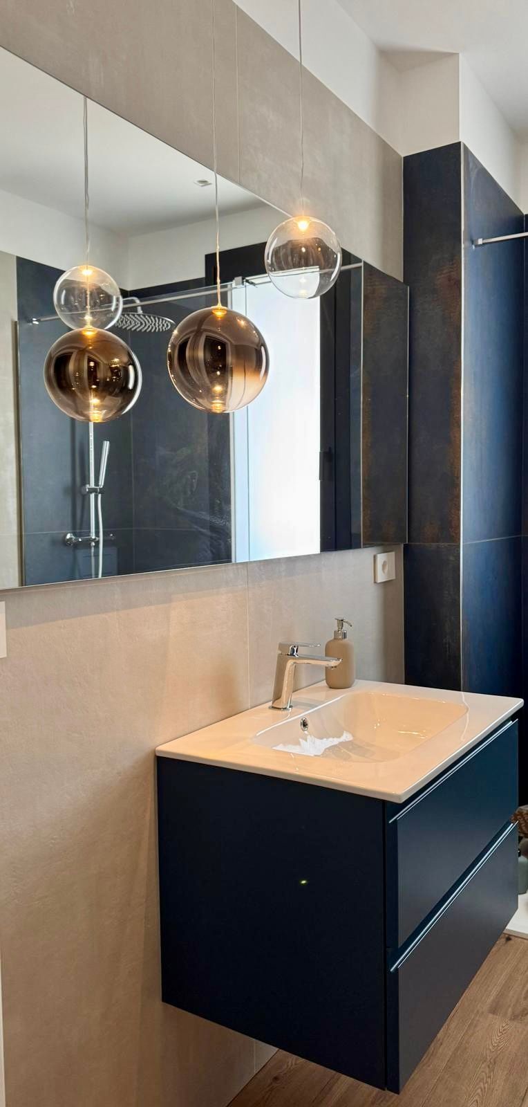 Bagno con mobile lavabo blu scuro, lampade a sospensione dorate e grande specchio.