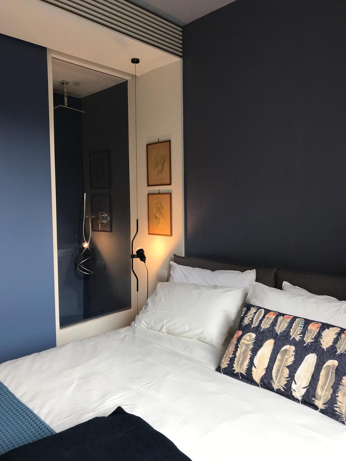 Angolo della camera da letto con pareti blu scuro e bianche, letto con lenzuola bianche, cuscino a fantasia e una lunga lampada a sospensione.