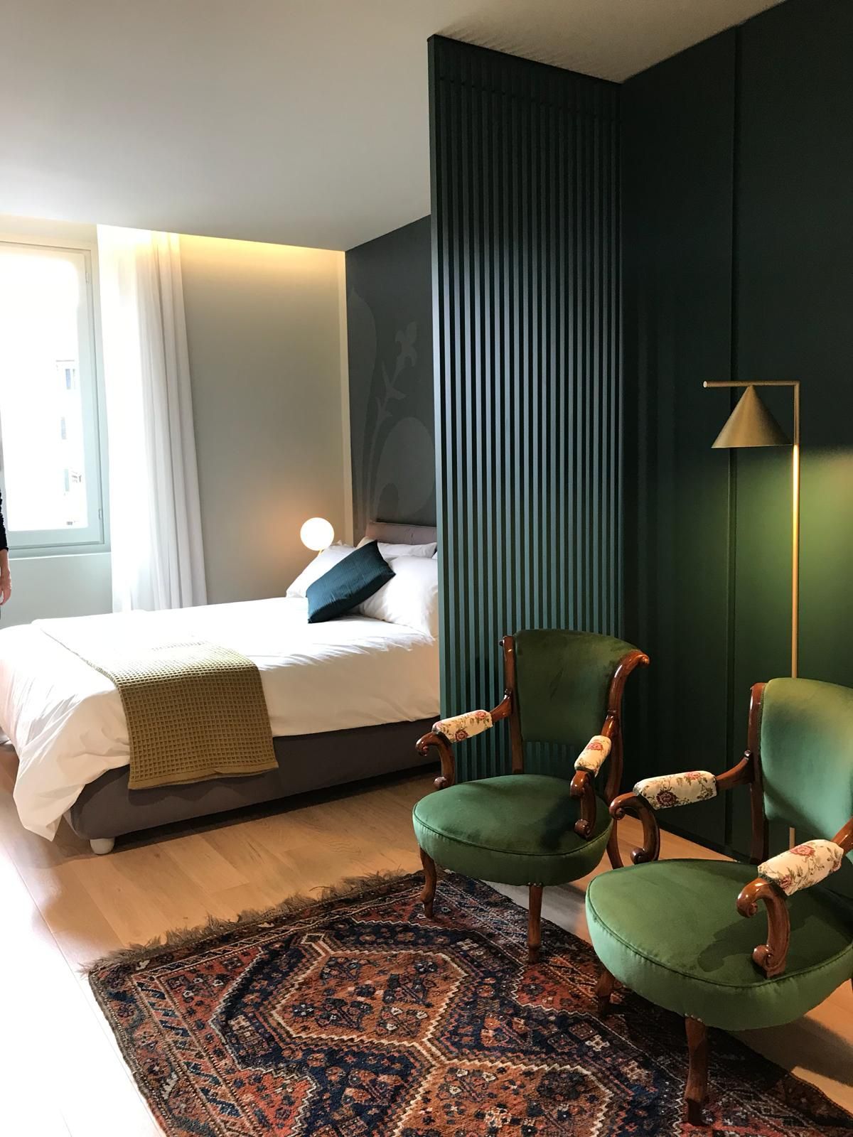Camera da letto con parete verde, due poltrone verdi, letto con lenzuola bianche e tappeto a fantasia.