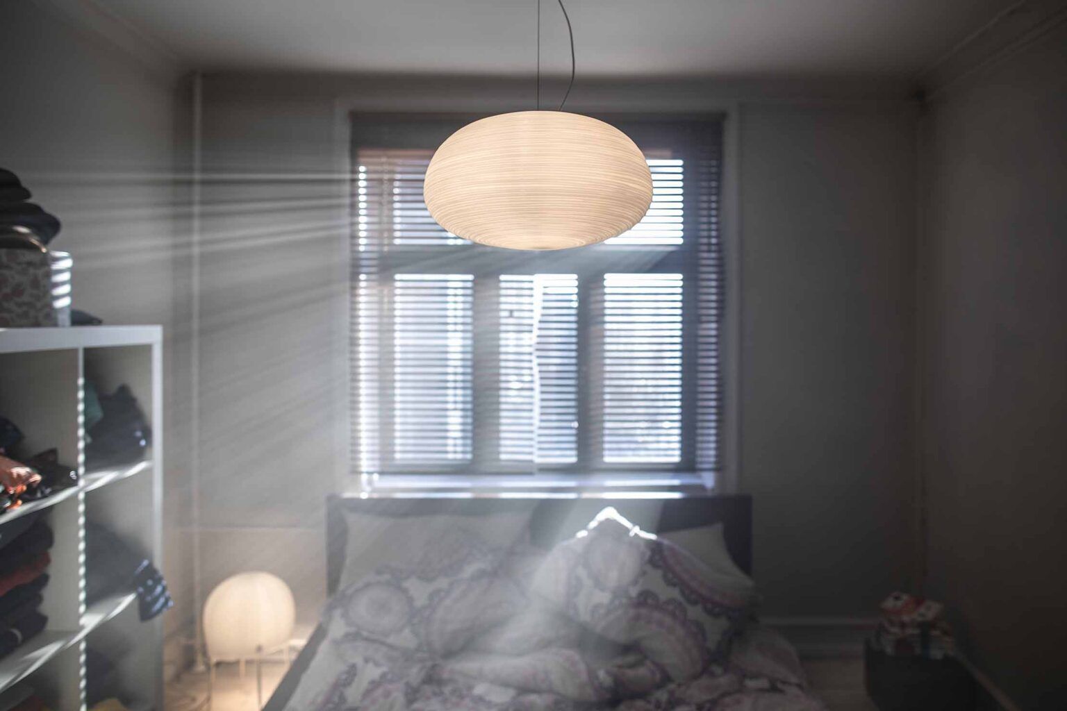 Interno della camera da letto con la luce del sole che filtra attraverso una finestra, illuminando un letto e una lampada decorativa sospesa.