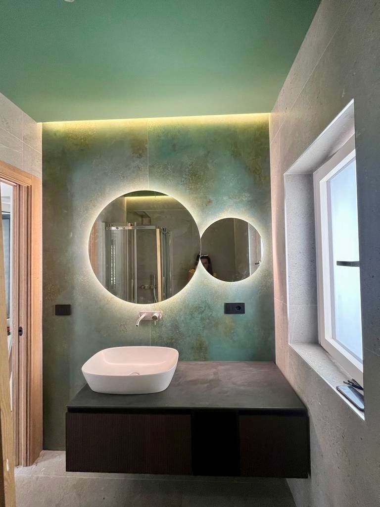 Bagno moderno con specchio rotondo illuminato, lavandino bianco e mobiletto sospeso scuro. Parete verde e grigia.