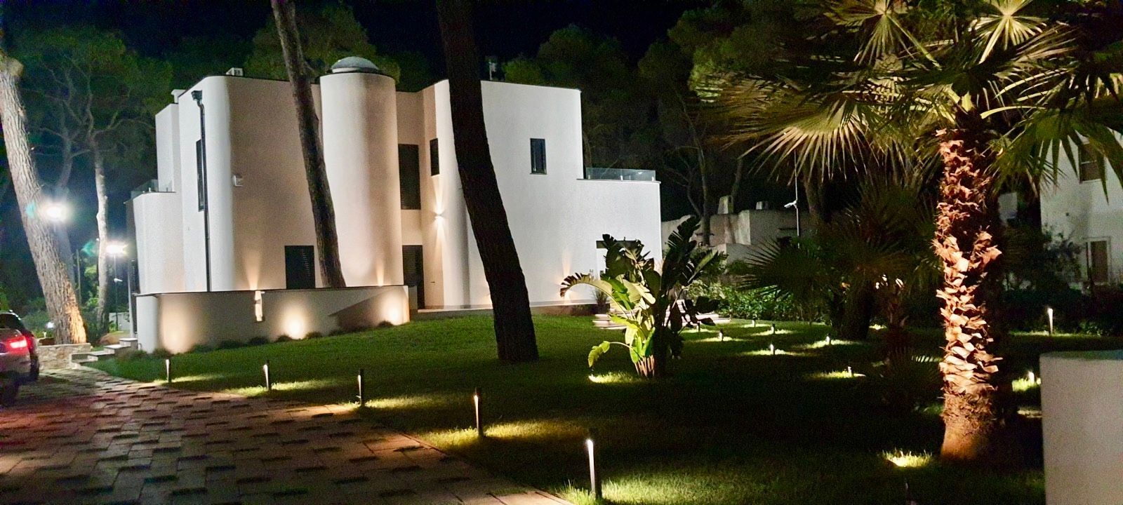 Vista notturna di un edificio bianco con luci esterne, alberi e una palma.