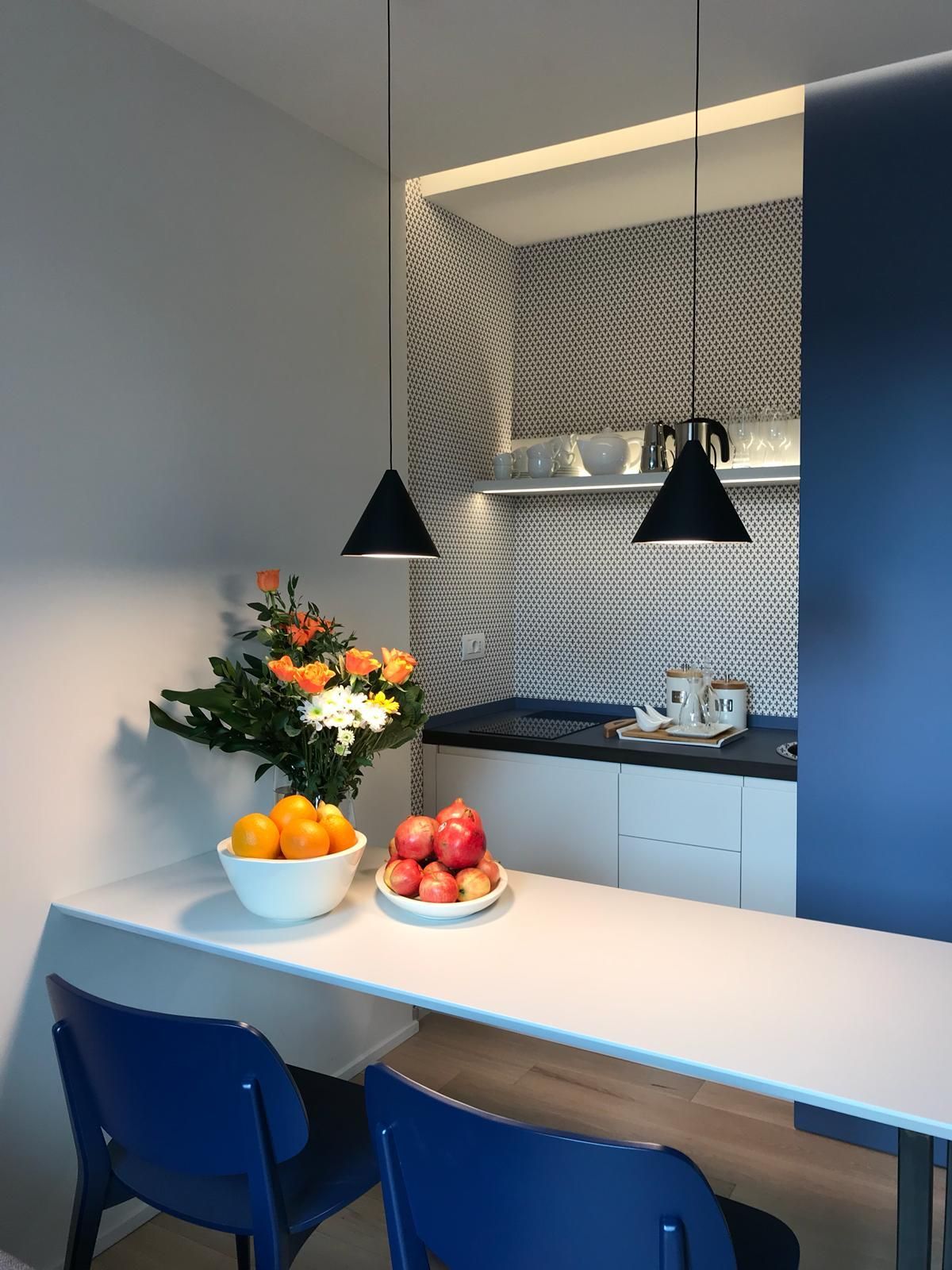 Un angolo cottura moderno con piano di lavoro bianco, sedie blu, lampade a sospensione e fruttiere.