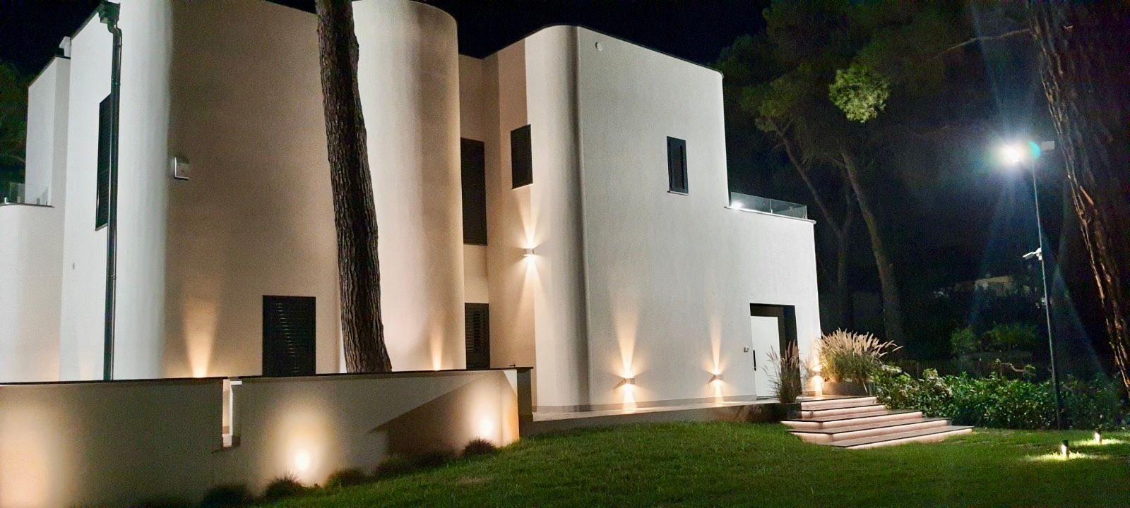 Un moderno edificio bianco di notte con illuminazione esterna. Prato e alberi di fronte.