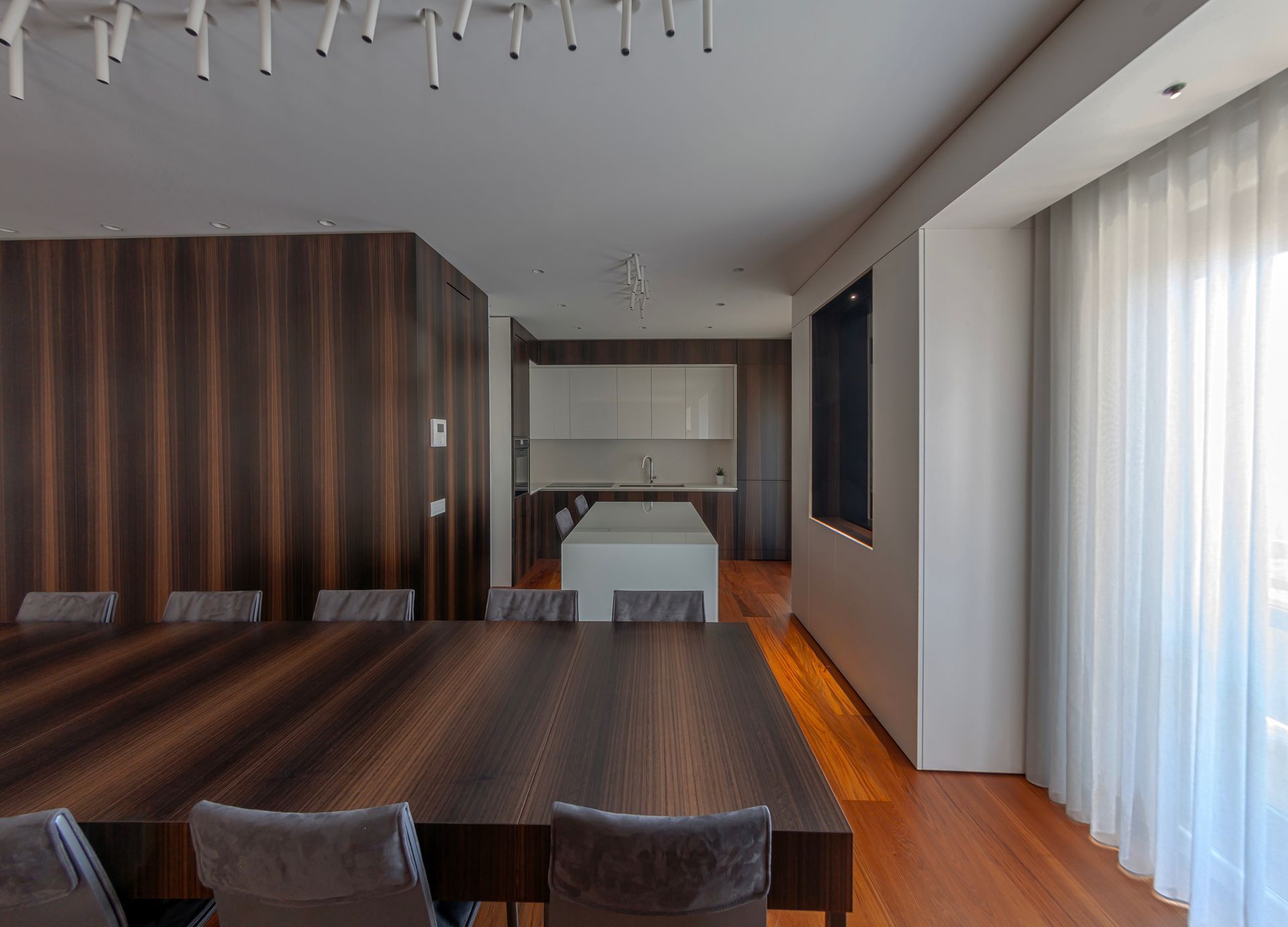 Sala da pranzo con un lungo tavolo in legno, una parete rivestita in legno scuro e una vista sulla cucina.