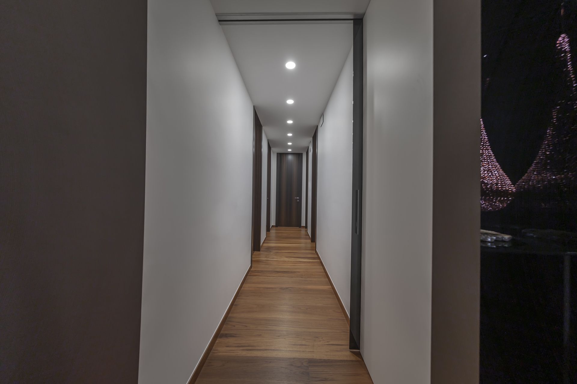 Corridoio stretto con pavimento in legno e pareti bianche, illuminato da luci da incasso nel soffitto.