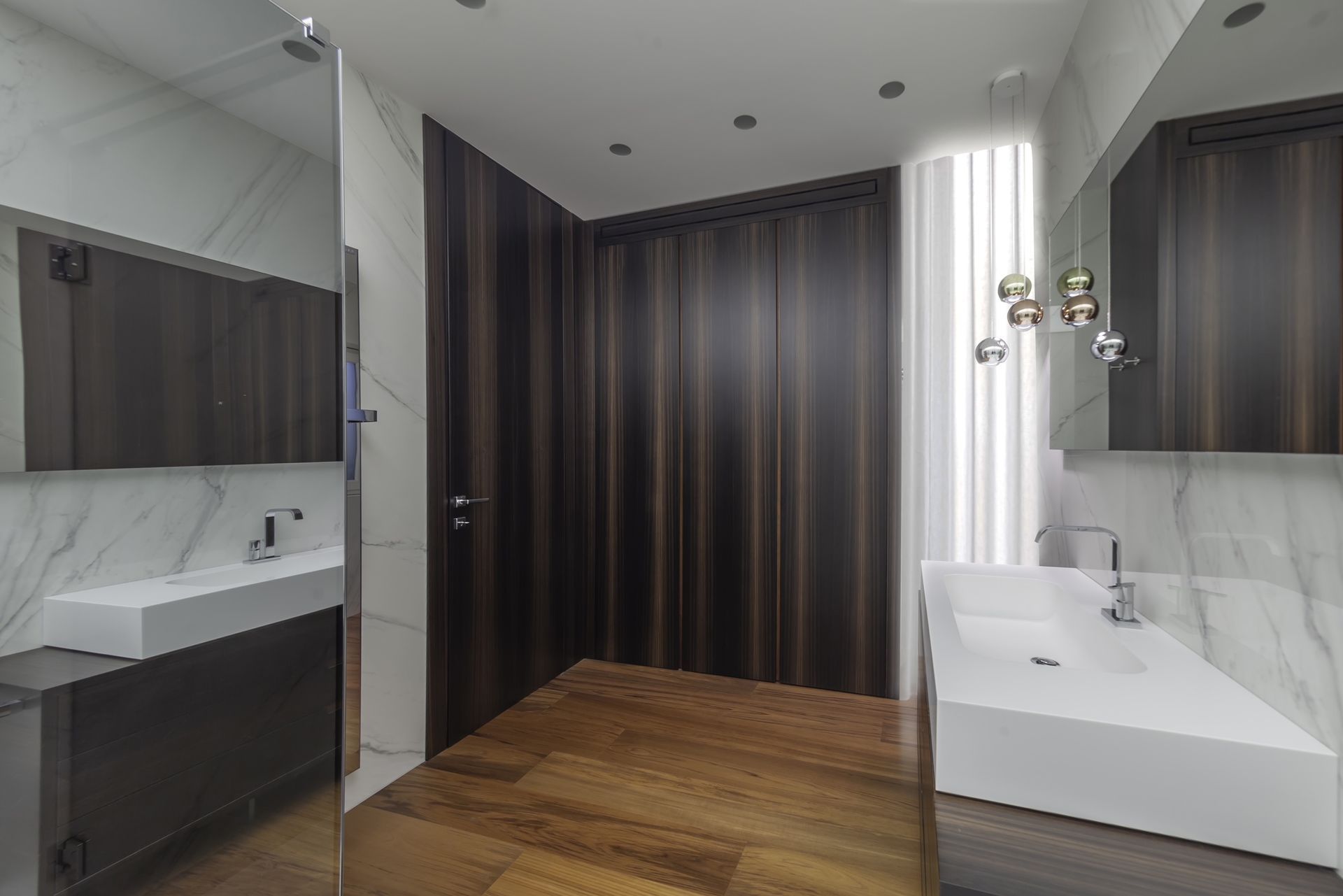 Bagno moderno con porta e pavimento in legno, lavandini bianchi, pareti in marmo.