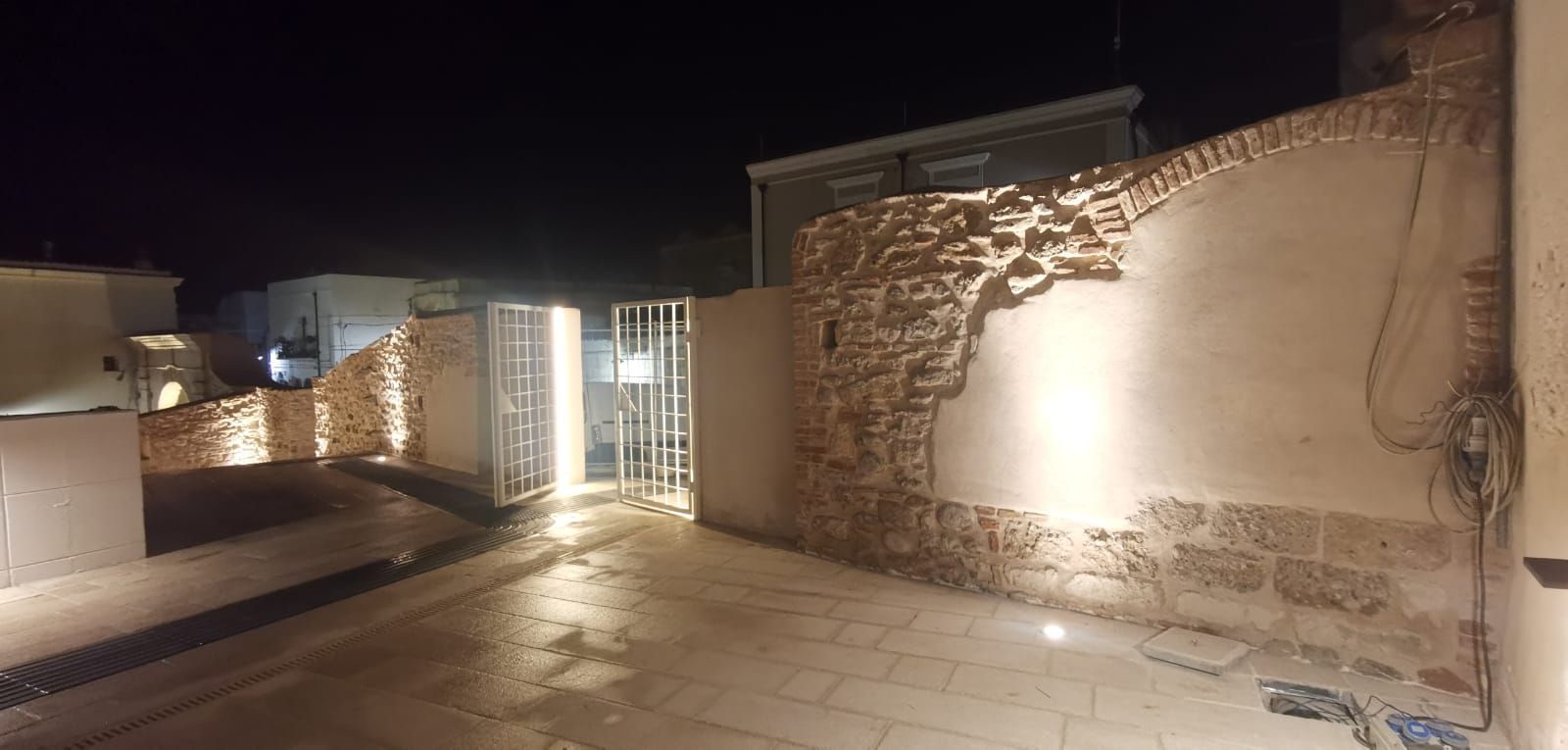 Vista notturna di uno spazio esterno con muri in pietra, illuminazione e un cancello.