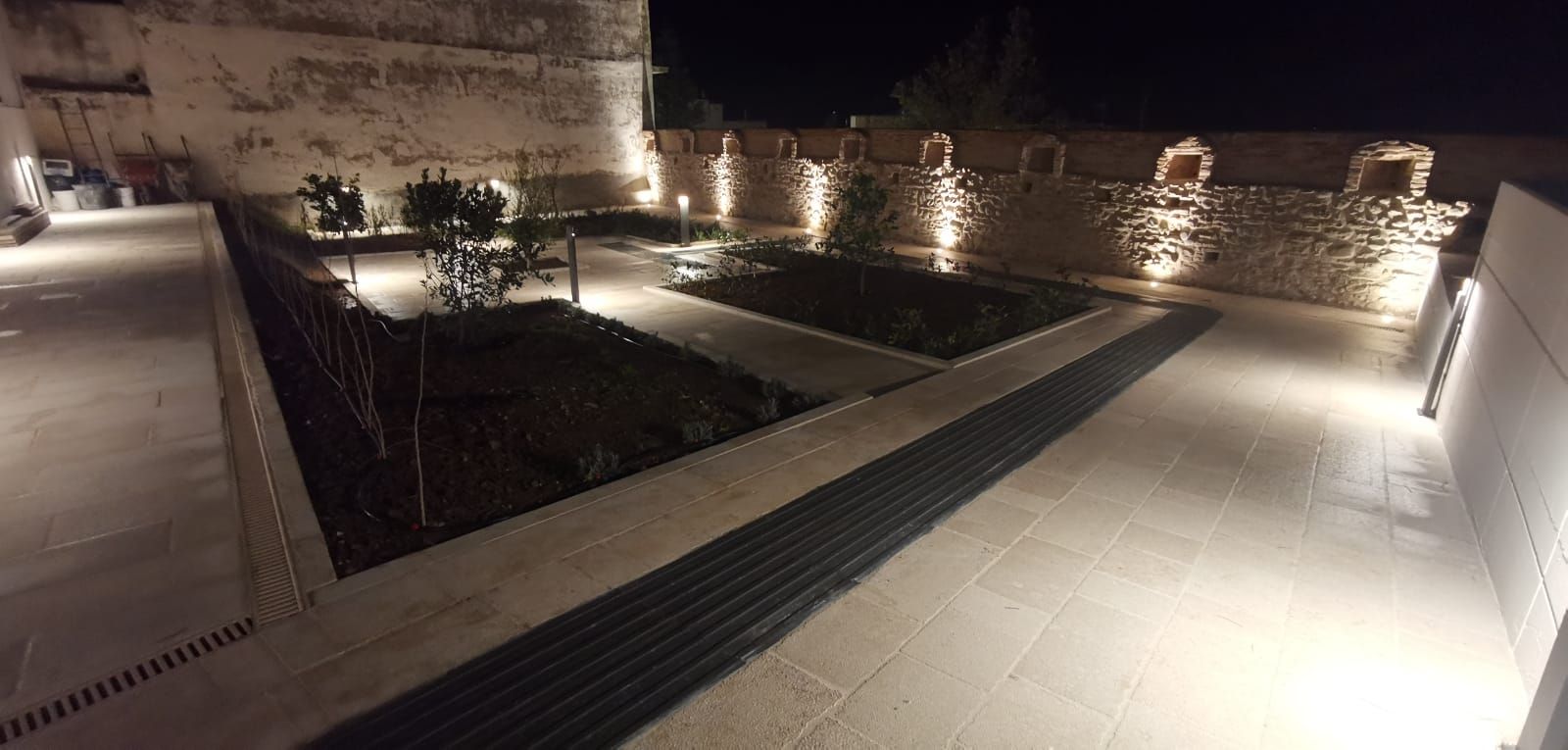 Vista notturna di uno spazio esterno illuminato con muri in pietra, piante e un sentiero illuminato.