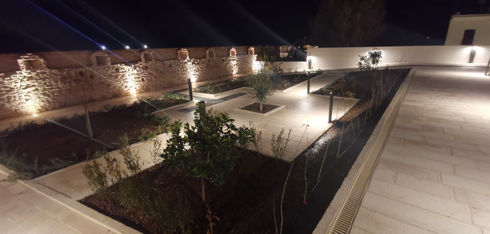 Vista notturna di un cortile con giardino, muri in pietra, varie piante e vialetti illuminati.