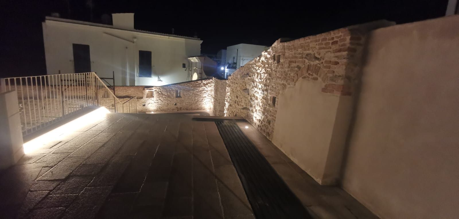 Uno scatto notturno di un muro di mattoni e intonaco con luci calde. Passerella in legno e edificio sullo sfondo.