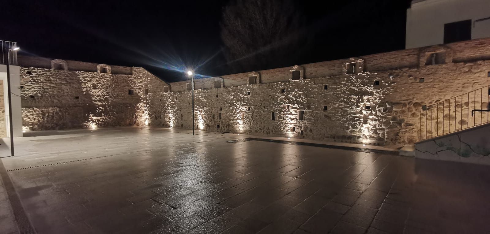 Vista notturna di un muro di pietra illuminato da faretti, che si riflette su una superficie bagnata.