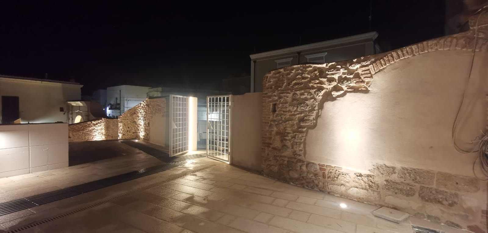 Vista notturna di un cortile con muri in pietra e un cancello metallico illuminato da faretti.