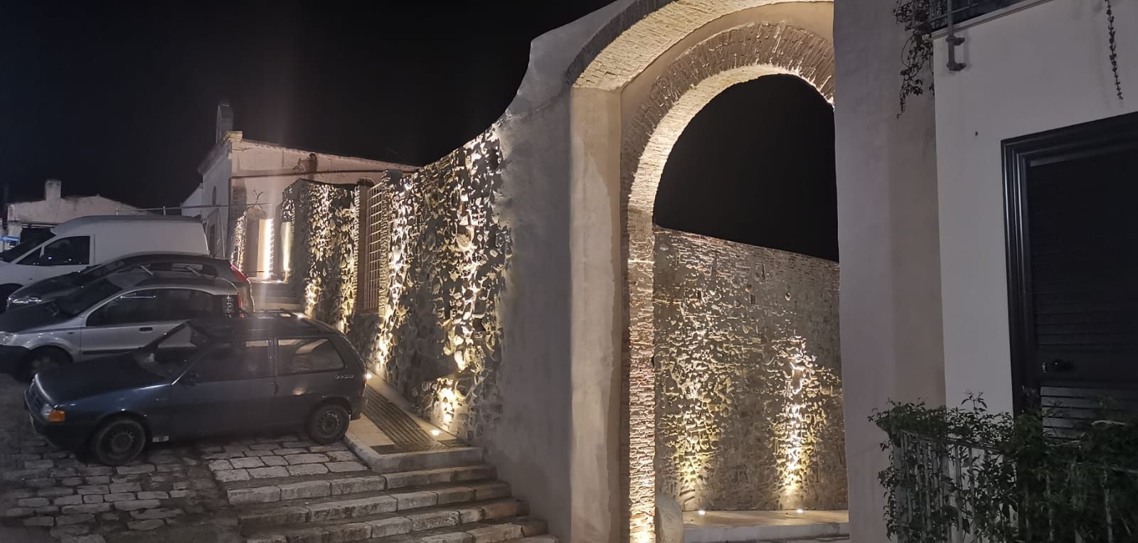 Esterno notturno con arco in pietra illuminato, gradini e auto parcheggiate.